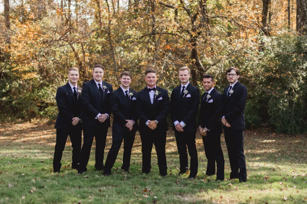 A wedding party of seven men in black formal attire poses against the vibrant autumnal foliage of our outdoor grounds at Lemon Ridge Events in Jasper, Alabama. This image highlights one of our versatile outdoor photography and ceremony spaces, providing a natural and sophisticated backdrop for milestone celebrations. Whether hosting an intimate micro-wedding or a full-scale celebration, our venue's expansive facility and scenic wooded areas offer the perfect setting for effortless and personal wedding photography.