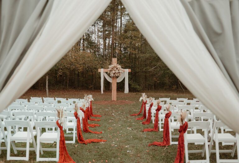 This image showcases one of our versatile outdoor ceremony locations at Lemon Ridge Events. Framed by elegant white draping, the scene features a large wooden cross altar adorned with pampas grass and neutral fabric. Set against our lush Jasper woodland backdrop, the arrangement includes white folding chairs accented with terracotta fabric and dried florals along the aisle. This setup highlights our ability to provide a beautiful, personalized setting for weddings and elopements, ensuring a stunning and effortless backdrop for our couples’ special day.