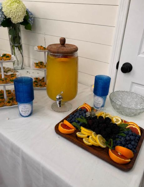 A catered baby shower refreshment station featuring a large glass beverage dispenser filled with orange punch, accompanied by stacks of blue custom-labeled cups. Beside the drinks sits a wooden serving platter elegantly arranged with fresh blueberries, blackberries, and citrus slices, next to a bowl of ice and a tiered stand of savory hors d'oeuvres. The clean, white backdrop creates an inviting and professional aesthetic for our event hosting services.
