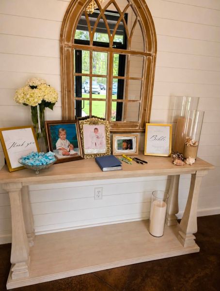 A beautifully styled baptism welcome table featuring framed portraits, a guest book, a Bible, 'thank you' signage, and fresh white hydrangeas, set against a rustic shiplap wall and arched window.