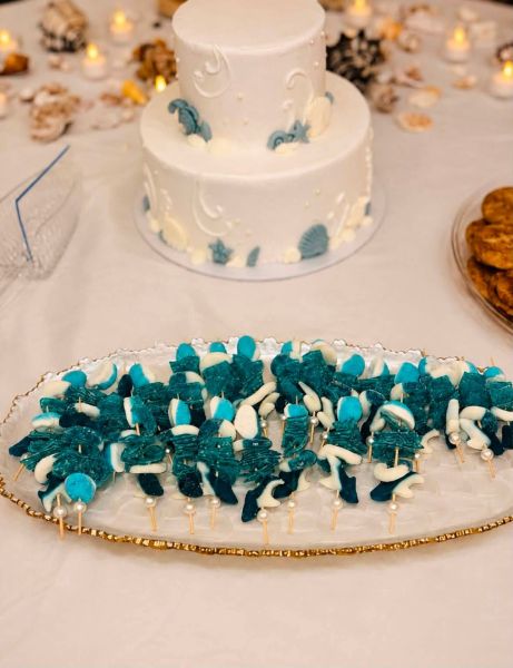A two-tiered white cake decorated with edible seashells and sea-inspired accents, centerpiece to a table setting. In the foreground, a decorative platter holds a series of blue and white gummy candy skewers, themed to match the ocean aesthetic. The table is scattered with small shells and tea light candles, creating a cohesive coastal party atmosphere perfect for a themed event.