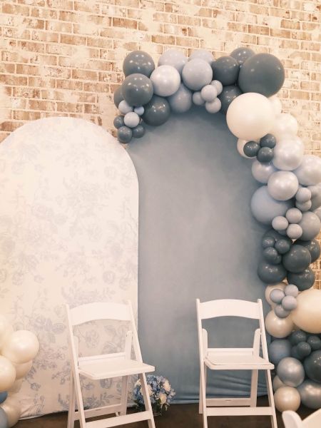 This setup features a sophisticated layered backdrop design, combining a floral-patterned white arch panel with a solid dusty blue velvet arch. We have accented this with a modern, organic balloon garland in varying shades of slate, navy, soft blue, and crisp white. Two white folding chairs are positioned in front, creating a perfect photo opportunity or seating area for events like baby showers, bridal reveals, or birthdays. The exposed brick background adds a rustic, high-end texture to this elegant display.