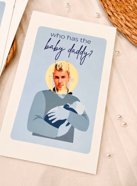 A light blue baby shower game card titled 'Who has the baby daddy?' with a circular gold scratch-off area that reveals a picture of Justin Bieber.