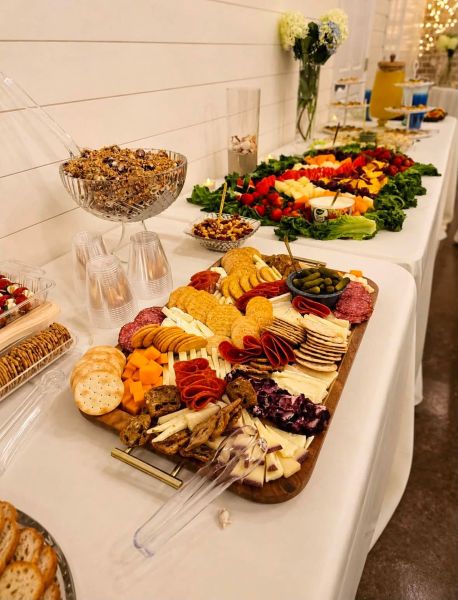 A high-angle shot of a beautifully curated charcuterie board featuring an assortment of crackers, sliced cheeses, cured meats, and pickles, displayed on a wooden platter. In the background, an elongated buffet table is laden with fresh fruit platters, bowls of granola, and additional hors d'oeuvres, set in a bright, modern event space. This image captures our commitment to elegant presentation and high-quality catering services, ideal for showcasing our ability to provide sophisticated refreshments for weddings, corporate gatherings, and special celebrations.