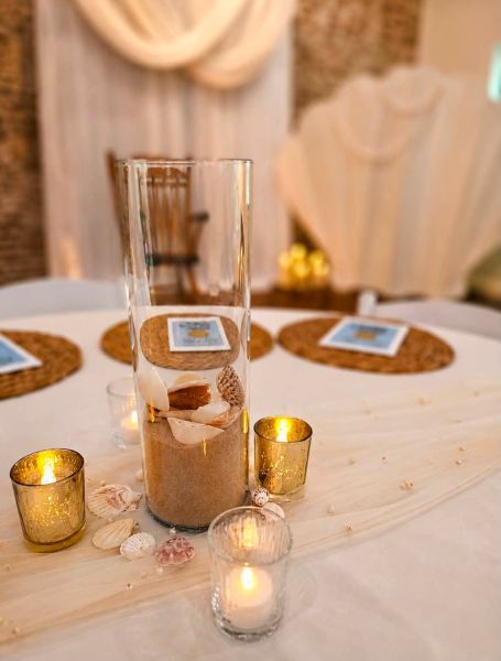 A close-up of a beach-themed table centerpiece designed for an event. It features a tall glass cylinder filled with sand and various seashells, surrounded by shimmering gold votive candles and scatter pearls on a draped fabric runner. Woven placemats are visible in the background, set for a relaxed, elegant gathering. This setup captures our signature approach to creating intimate, coastal-inspired table arrangements that bring the warmth and beauty of the shore to any indoor venue.