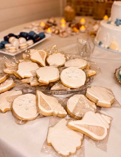 A close-up view of our custom-decorated sugar cookies featuring intricate seashell and sand dollar designs. These hand-iced treats are individually wrapped and displayed on a platter as part of a wedding dessert table. The neutral, elegant aesthetic complements the surrounding beach-themed decor, including blue-tinted macarons and a tiered celebration cake in the background. Perfect for couples looking to add a personalized, thematic touch to their coastal wedding reception.