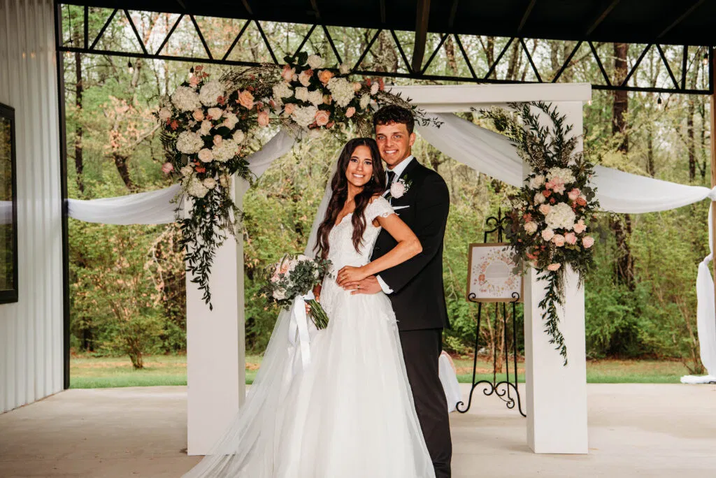 A newlywed couple poses in front of our elegant white ceremony arbor, showcasing one of our versatile outdoor-covered spaces at Lemon Ridge Events. The arch is beautifully adorned with lush floral arrangements in soft peach, pink, and white. The backdrop features our serene, wooded landscape, providing a picturesque and weather-protected setting for weddings and elopements. This captures the effortless beauty and personal touch we provide to every couple celebrating their milestone in our flexible Jasper venue.