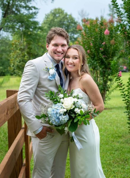 A portrait of a radiant bride and groom standing together outdoors beside a wooden rustic fence. The bride holds a lush floral bouquet featuring blue hydrangeas and white roses, which match the groom's boutonniere. Both are smiling warmly at the camera, highlighting the natural beauty and joyful atmosphere of their outdoor summer wedding. This image captures the classic, timeless feel we aim for in all our wedding portrait sessions.