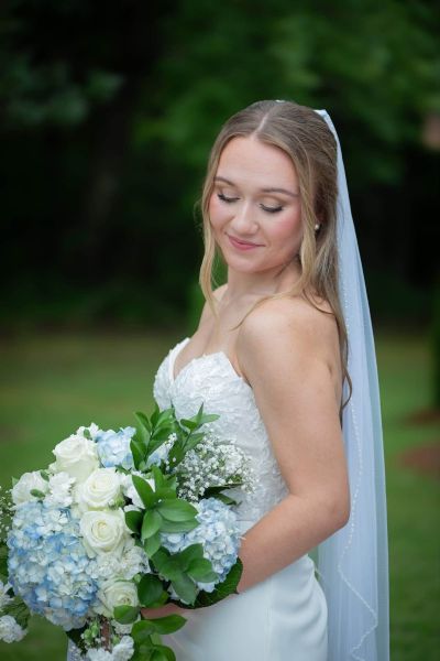 A bride in a sleek white strapless gown stands on a patio holding a bouquet of blue hydrangeas and white roses, with a rustic wooden wedding cross decorated with matching flowers in the background.