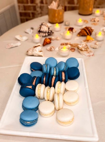 A rectangular serving platter featuring an assortment of blue and white macarons, presented as the centerpiece for a coastal-themed event. The cookies are arranged in rows, complementing a table backdrop decorated with seashells and warm, glowing tea light candles. This setup highlights our ability to tailor dessert presentations to match specific event aesthetics and color palettes.