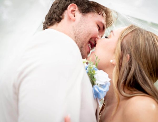 A close-up, intimate portrait of a bride and groom sharing a tender moment under a clear umbrella. The soft, bright lighting captures the joy on their faces, shielded from the elements. This image perfectly illustrates how we embrace the unique, unpredictable moments of a wedding day, turning rainy weather into a romantic and memorable part of the couple's special celebration.
