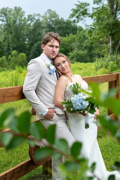 A bride and groom pose intimately against a rustic wooden fence at our venue. The bride wears a strapless white gown and holds a bouquet featuring blue hydrangeas, while the groom wears a tan suit with a matching blue floral boutonniere. They are surrounded by the lush green foliage of our outdoor property, showcasing the natural beauty of our grounds as a backdrop for wedding photography.