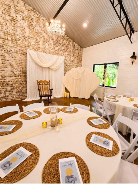 A baby shower setup featuring a shell-shaped backdrop, a wooden rocking chair, and tables decorated with neutral linens and game cards against an exposed brick wall.