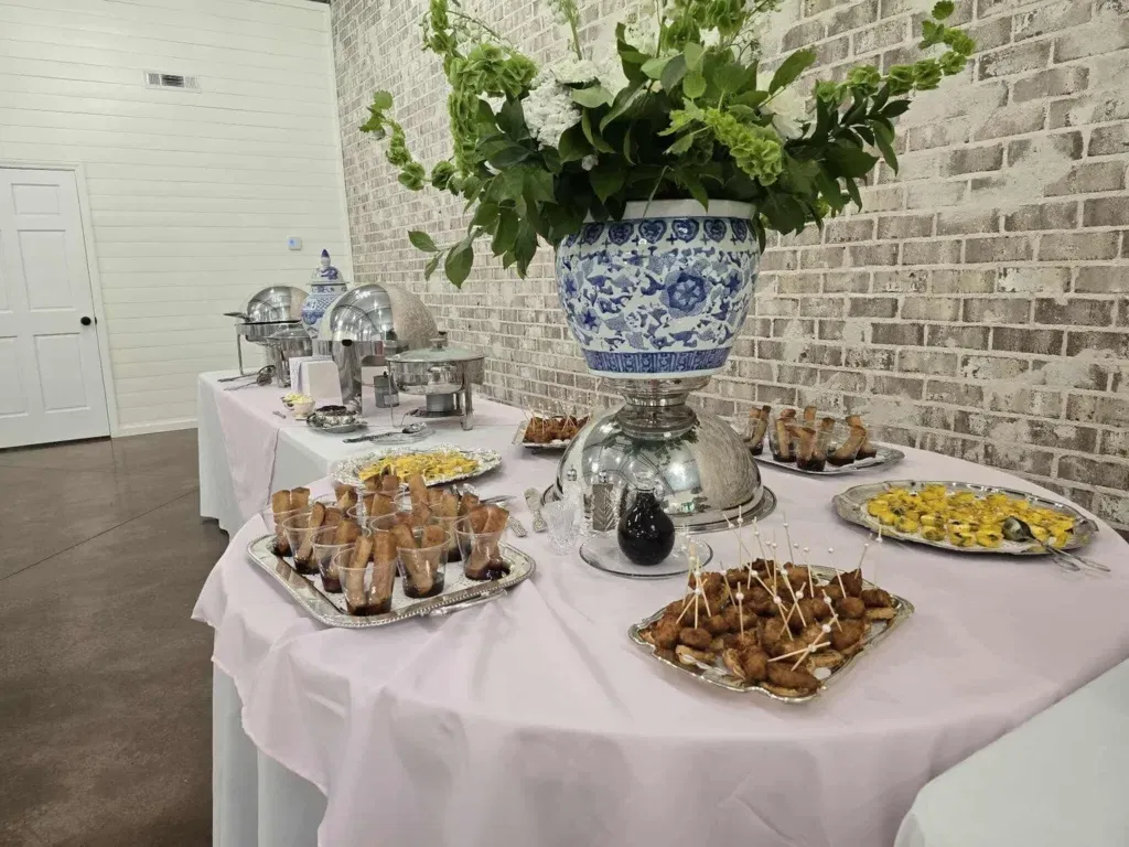 A sophisticated indoor catering display at Lemon Ridge Events, featuring an array of hors d'oeuvres and polished silver chafing dishes. The setup is anchored by a large, classic blue and white chinoiserie floral arrangement against our signature whitewashed brick wall. This scene highlights the versatility of our 10,000-square-foot facility, demonstrating how we accommodate professional catering with style. Our spacious interior provides a clean, elegant aesthetic, ensuring that every wedding, shower, or party benefits from a beautiful and functional food service area.