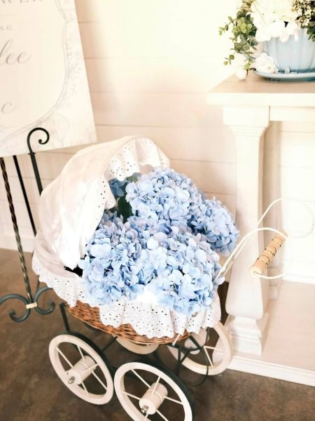 A vintage-style wicker baby carriage styled as a decorative floral centerpiece. The pram is filled with voluminous light blue hydrangea blossoms and accented with white eyelet lace trim. It sits in a bright, rustic-themed event space next to a white wooden pedestal and a welcome sign, perfect for showcasing our baby shower decor styling services.
