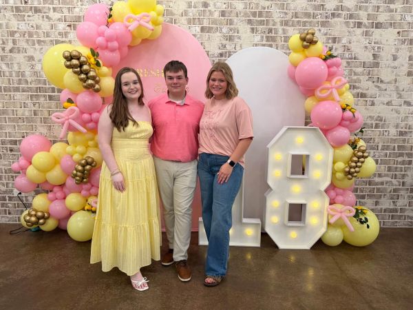 A group of three young adults posing in front of a professional custom 18th birthday balloon display featuring a double-arch backdrop, pink and yellow balloon garland with lemon motifs, and a large illuminated '18' marquee.
