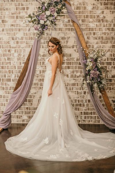A bride stands gracefully in front of our signature wooden triangular wedding arch, set against a rustic whitewashed brick wall. The arch is elegantly adorned with soft lavender fabric draping and vibrant floral arrangements in shades of purple and white. This image highlights our venue's versatile indoor ceremony backdrop, showcasing how our décor creates a romantic, polished, and intimate atmosphere for wedding photography.