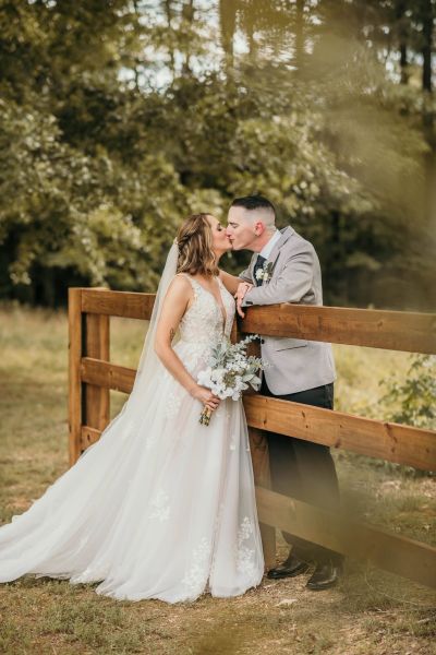 A tender portrait of a bride and groom sharing a kiss while leaning against a polished wooden fence on our property. The bride, wearing an elegant lace gown and veil, holds a floral bouquet, while the groom looks sharp in a gray blazer. This shot perfectly captures the intimate, natural beauty of our outdoor wedding venue, highlighting the soft, scenic greenery of the surrounding woods as a romantic backdrop for our couples.