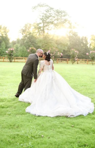A wide shot of a bride in an elaborate white lace gown and a groom in a dark suit sharing a romantic kiss in the center of our expansive, lush green event lawn. The late afternoon sun creates a warm, glowing lens flare behind them, highlighting our signature manicured grounds and the peaceful, private atmosphere we provide for wedding portraits.