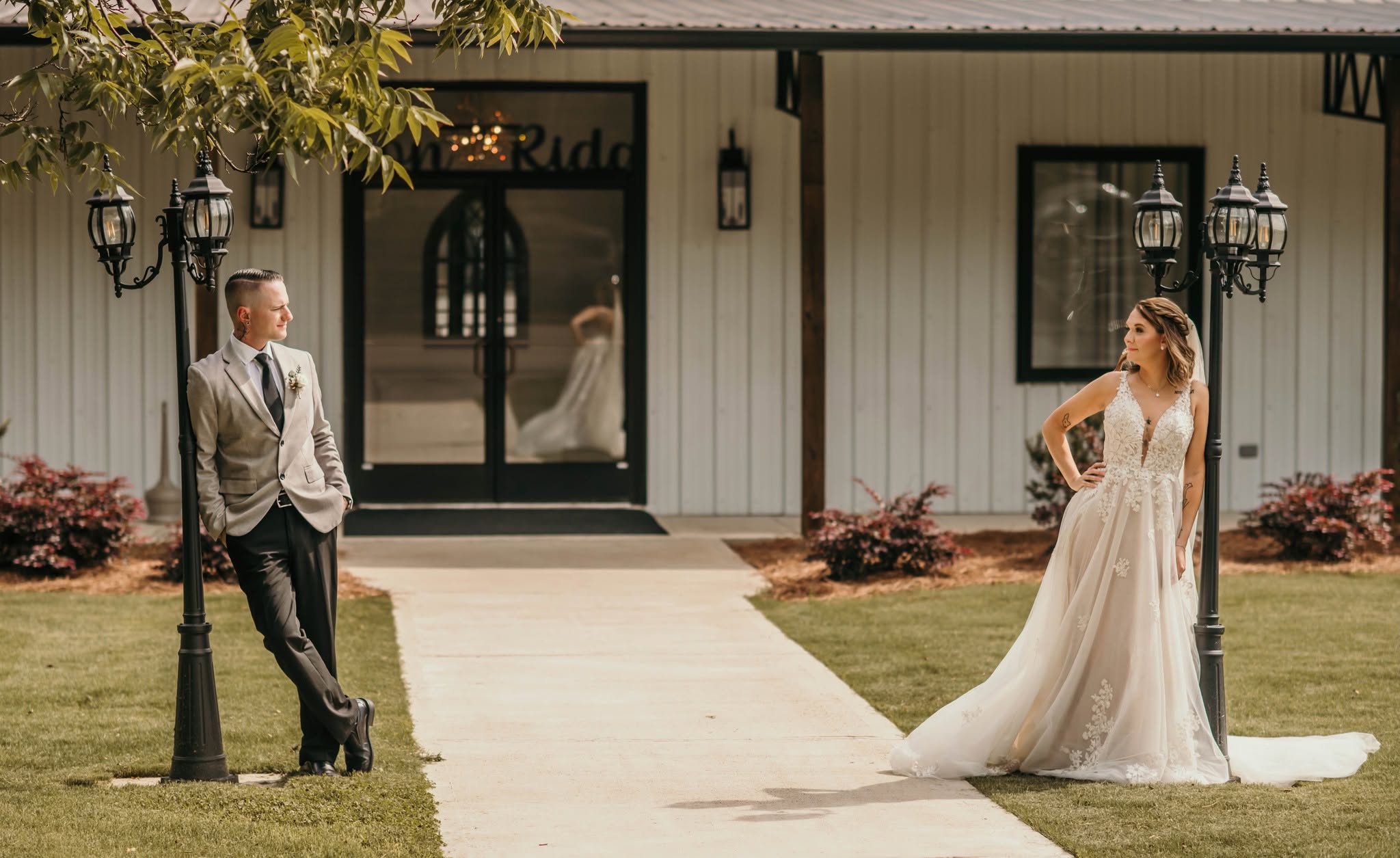 A wide shot capturing a bride and groom posing elegantly outside the main barn entrance. The groom leans against a vintage-style lamp post on the left, while the bride stands gracefully by a matching lamp post on the right. The white board-and-batten siding and black trim of the venue provide a crisp, modern-rustic backdrop, highlighting the beauty of our exterior grounds during a couple's wedding day portrait session.