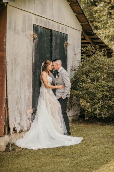 A tender portrait of a bride and groom in an intimate embrace, leaning against the rustic, weathered wooden doors of a historic barn. The scene highlights the contrast between the bride's elegant lace gown and the textured, natural backdrop, capturing a timeless, romantic wedding moment.
