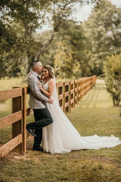 A tender moment featuring a bride and groom posing against a signature wooden rail fence. The groom is gently kissing the bride’s forehead while they lean against the rustic outdoor backdrop of the wedding venue grounds.