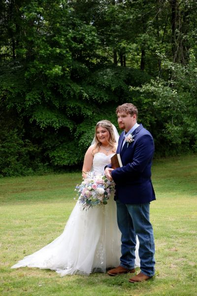 A bride in a white strapless gown and a groom in a navy blazer and jeans standing together on the grass at an outdoor wedding venue. The groom holds a Bible, and the bride carries a colorful bouquet.