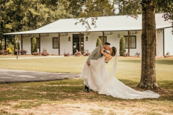 A romantic shot of a bride and groom sharing a dip and a kiss on our manicured front lawn. The image captures the couple in front of our signature white farmhouse, highlighting the venue's classic, rustic elegance as a backdrop for timeless wedding portraits. The sweeping train of the bride's gown and the lush greenery provide a perfect, natural setting for intimate post-ceremony photos.