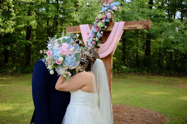 A bride and groom share a moment behind an oversized pink and blue bridal bouquet in front of a floral-adorned wooden wedding cross in a forested outdoor setting.