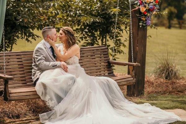 A bride and groom share an intimate, tender moment while sitting together on our venue’s rustic wooden porch swing. The couple is positioned in a loving embrace, highlighting the serene and romantic atmosphere of our outdoor gardens. The scene features our signature floral-adorned swing, serving as a perfect, picturesque backdrop for wedding portraits that capture the quiet joy of a newly married couple on their special day at our location.