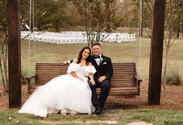 A bride in a white gown and a groom in a black tuxedo sitting together on a rustic wooden porch swing, with an outdoor wedding ceremony set up in the background.