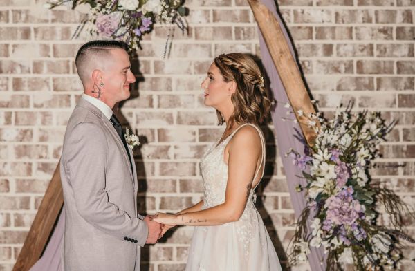 A bride and groom stand face-to-face, holding hands during their wedding ceremony at our venue. They are positioned in front of our signature wooden triangular arbor, which is elegantly decorated with lavender and white florals and soft purple drapery. The rustic white-washed brick backdrop provides a warm, intimate atmosphere for the couple as they share their vows. This shot captures the romantic essence of our indoor ceremony space, highlighting how beautifully our versatile decor complements a couple's special day.