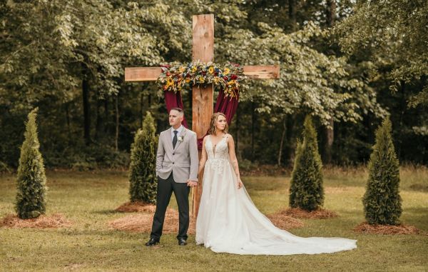A bride and groom stand hand-in-hand before our signature wooden cross altar. The structure is decorated with seasonal floral arrangements and deep burgundy fabric, set against a lush, wooded backdrop. This image highlights our venue's dedicated ceremony space, showcasing how our natural landscape and custom altar provide a beautiful, serene setting for couples to exchange their vows.