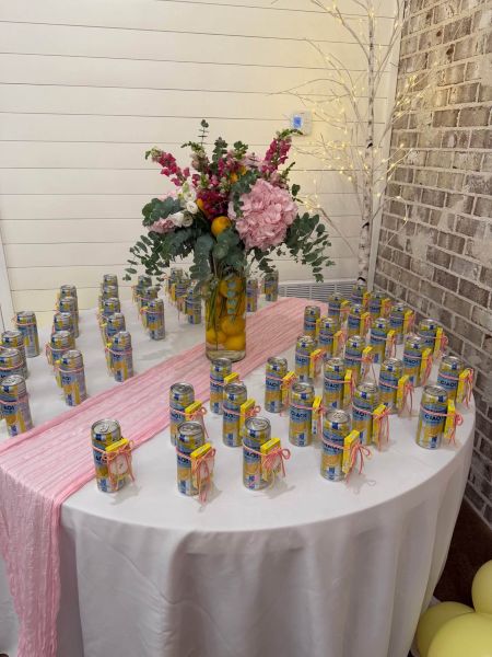 A round event table serving as a favor station, decorated with a pink runner, a lemon-filled floral centerpiece, and rows of Sanpellegrino Ciao lemon beverage cans with decorative tags.