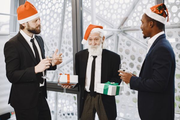 A group of diverse professional colleagues, including an older gentleman and younger associates, wearing Santa hats during an office holiday celebration. The team is holding gifts and glasses of champagne, reflecting the festive spirit and camaraderie within our professional environment. This image captures the balance of business networking and seasonal festivities, showcasing our workplace culture during the holiday season.