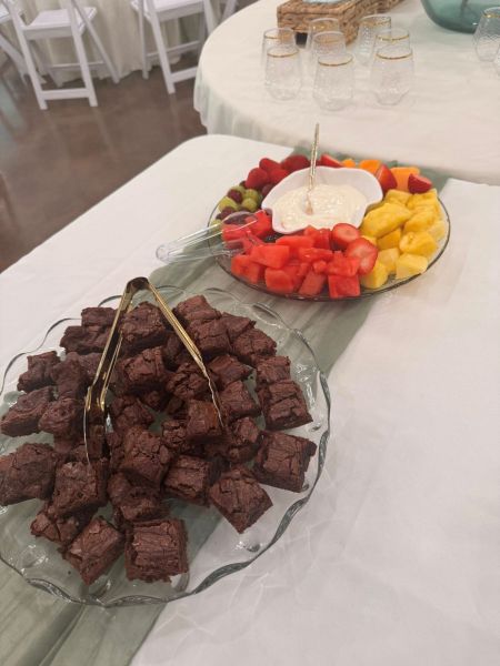 A catered display featuring a glass platter of bite-sized chocolate brownie squares alongside a fresh fruit platter with strawberries, watermelon, pineapple, and grapes served with a side of cream dip. Perfect for event refreshment stations, these platters offer a balanced mix of indulgent treats and light, healthy snacks for our guests.