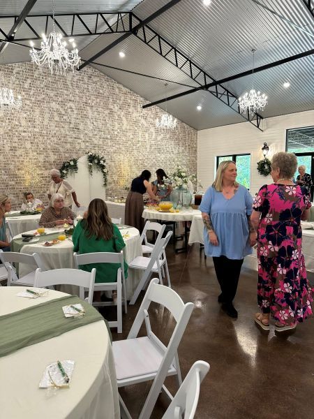 A view inside an event venue during a daytime social gathering, featuring round tables with white linens, sage green runners, and white folding chairs. The room has a rustic-chic aesthetic with a white-washed brick accent wall, vaulted ceilings, and crystal chandeliers.