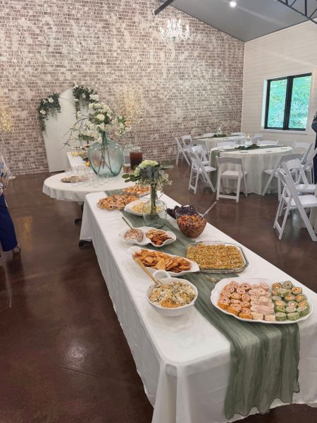 This photo showcases our main dining hall set up for a catered event. The long buffet table is dressed with a sage green runner and features an assortment of appetizers, including pinwheels, dips, and chips. The background displays our signature whitewashed brick wall, an elegant floral-adorned backdrop, and rounds set with white chairs, providing a perfect example of how our venue accommodates sophisticated, full-service catering displays for weddings and private parties.