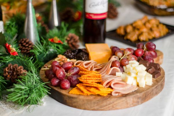 A rustic wooden platter featuring a curated selection of holiday appetizers, including sliced deli meats, cubed white cheddar, sharp cheddar slices, red grapes, crackers, and dark chocolate pieces. The board is elegantly presented on a table adorned with festive evergreen branches, pinecones, and red accents, set beside a bottle of red wine. This display highlights our signature holiday entertaining style, perfect for seasonal celebrations and upscale catering events.