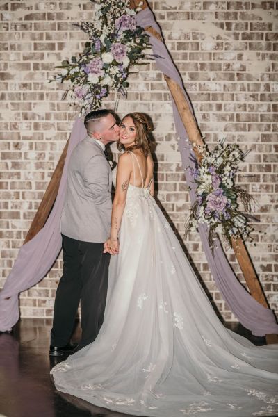 A romantic portrait of a bride and groom captured in front of our signature wooden triangular wedding arch. The arch is adorned with elegant lavender and white floral arrangements and soft purple fabric draping, set against our rustic whitewashed brick feature wall. This image highlights our venue's capability to provide a stylish, intimate, and professionally styled backdrop for wedding ceremonies.