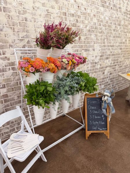 A white tiered metal stand displaying an assortment of fresh-cut flowers, foliage, and textured fillers in white buckets. Beside the stand, a wooden chalkboard sign invites guests to build their own custom bouquet by selecting three stems of greenery, three flowers, and three spiky fillers. White guest folding chairs and paper bouquet wraps are positioned nearby, ready for customers to create their personalized floral arrangements at our pop-up flower bar.
