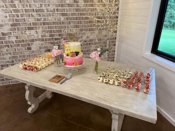 A vibrant, citrus-themed dessert display set on a rustic wooden table against a brick wall. The centerpiece is a dual-toned yellow and pink "Freshly Eighteen" birthday cake adorned with fresh flowers and citrus slices. Surrounding the cake are individual dessert shooters, including strawberry and cream parfaits and chocolate-layered treats. This setup highlights our ability to combine custom tiered cakes with elegant, grab-and-go finger desserts for milestone celebrations.
