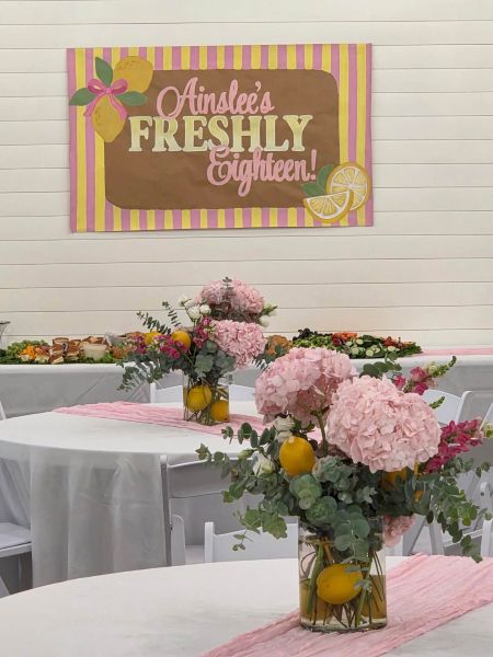 A vibrant, lemon-themed party setup for an eighteenth birthday celebration. The focal point is a custom banner reading "Ainsley’s FRESHLY Eighteen!" hung against a white shiplap wall. The tables are decorated with crisp white tablecloths, light pink runners, and elegant centerpieces featuring lush pink hydrangeas, greenery, and fresh lemons submerged in glass vases. The background includes a buffet table prepared with party snacks, highlighting our ability to coordinate fresh, thematic décor for milestones and special events.
