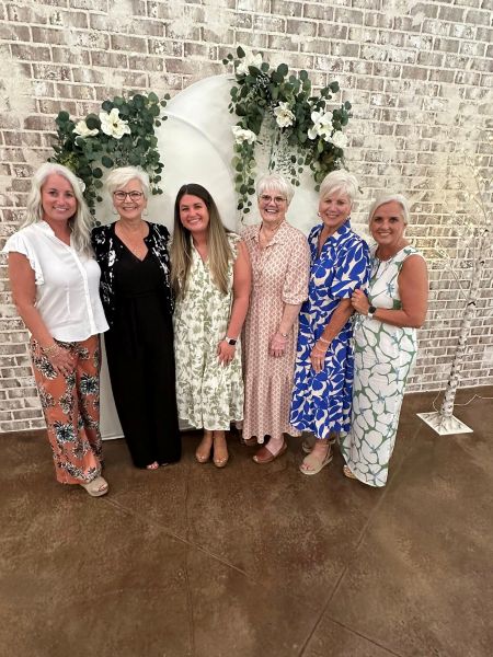 A group of six women smiling together in front of a white floral arch and rustic brick wall. This image captures the joyful, celebratory atmosphere of our venue, highlighting the perfect backdrop we provide for gatherings, reunions, and special occasions with friends and family.
