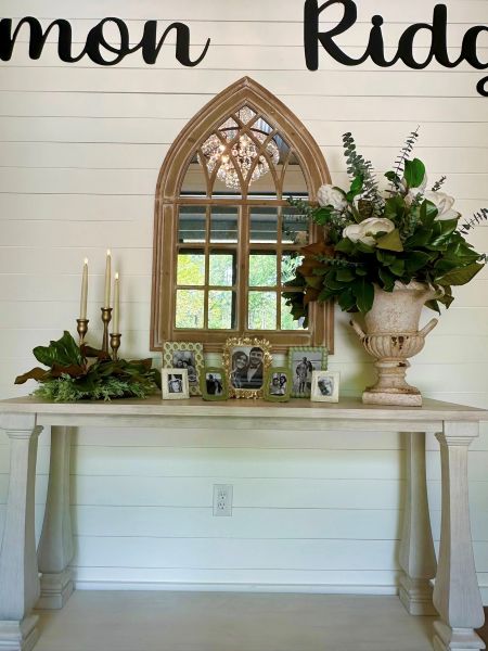 This vignette showcases our venue's elegant entryway decor, featuring a rustic wooden console table adorned with soft candlelight, a lush magnolia floral arrangement, and an arched mirror reflecting our signature chandelier. A collection of framed couple portraits adds a warm, personalized touch, highlighting the romantic atmosphere we cultivate for weddings and special events at Common Ridge.