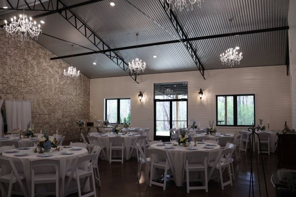 A wide shot of The Grand Hall event venue set for a reception, featuring high ceilings with chandeliers, exposed brick walls, and round tables with white linens.