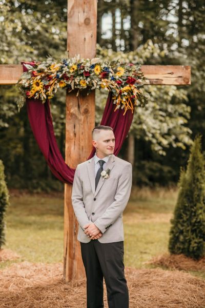 A portrait of a groom standing at our signature wooden cross altar during an outdoor ceremony. The altar is decorated with sunflowers, greenery, and draped burgundy fabric, showcasing our venue’s ability to customize ceremony decor. The groom is wearing a light grey suit jacket and dark trousers, posed in front of the natural, wooded background of our venue grounds. This image captures the intimate atmosphere and rustic elegance provided at Heritage Oaks.