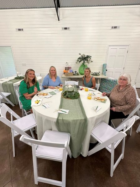 Four guests seated at a round table decorated with sage green runners, floral accents, and white chairs inside our event venue. The table is set for a casual luncheon or shower, showcasing our bright, neutral space and versatile, elegant event layout. The atmosphere is warm and inviting, perfect for intimate gatherings, workshops, or milestone celebrations hosted at our facility.