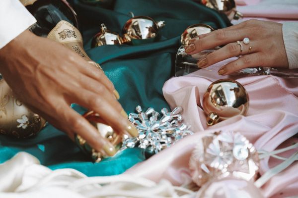 A close-up shot capturing the spirit of seasonal festivities. The image features a champagne bottle and sparkling ornaments arranged on luxurious teal and pink satin fabric. Two hands are visible, one adjusting a gold bauble and the other resting near a champagne flute, highlighting the sophisticated, celebratory atmosphere of our holiday events. The focus on textures and metallic accents reflects the premium experience we curate for our clients during the winter season.