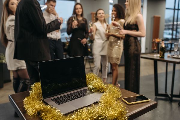 A laptop sits on a wooden desk decorated with festive gold tinsel in the foreground, with a group of professionals socializing and holding champagne glasses in the blurred background at a holiday party.